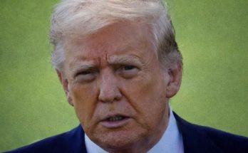 Close-up of a man with blond hair wearing a dark suit and red tie against a green