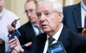 A man with white hair in a dark suit and striped tie speaks to reporters holding