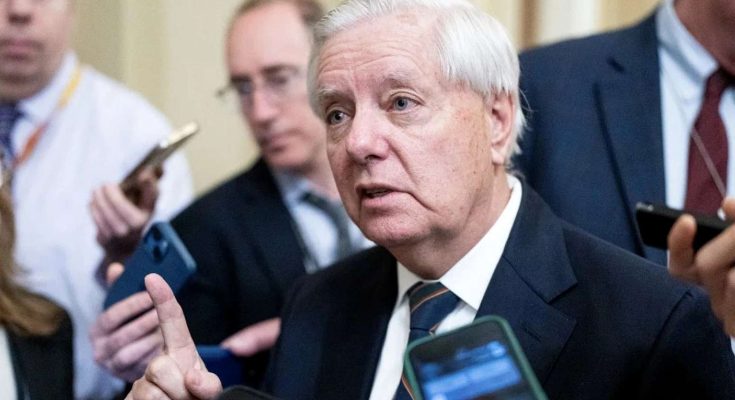 A man with white hair in a dark suit and striped tie speaks to reporters holding