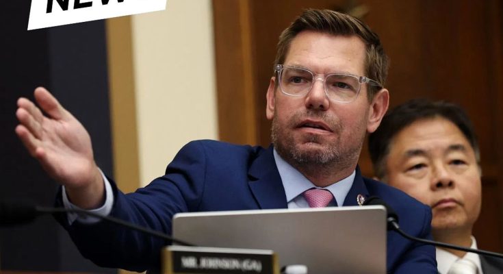 A man in a blue suit with a pink tie and clear glasses gestures with his right hand