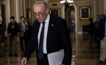Chuck Schumer walking through a Capitol hallway holding papers, wearing a dark suit and