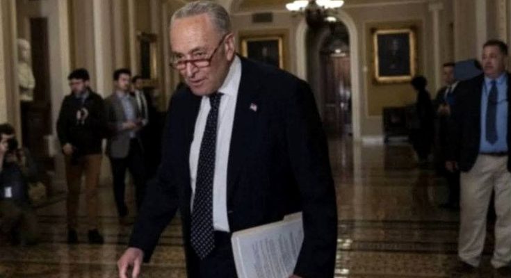Chuck Schumer walking through a Capitol hallway holding papers, wearing a dark suit and