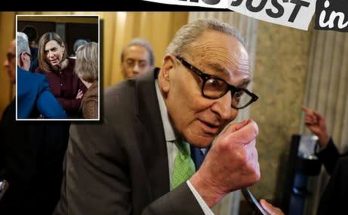 An older man in a suit and glasses is speaking while holding a phone, with a smaller