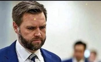 A man with brown hair and a beard wearing a blue suit, white shirt, and striped tie