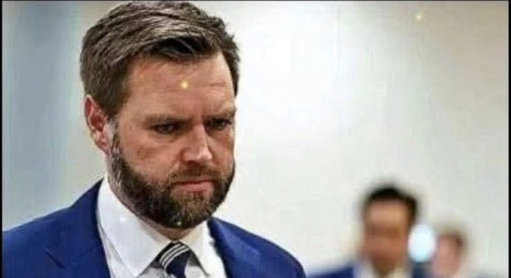 A man with brown hair and a beard wearing a blue suit, white shirt, and striped tie
